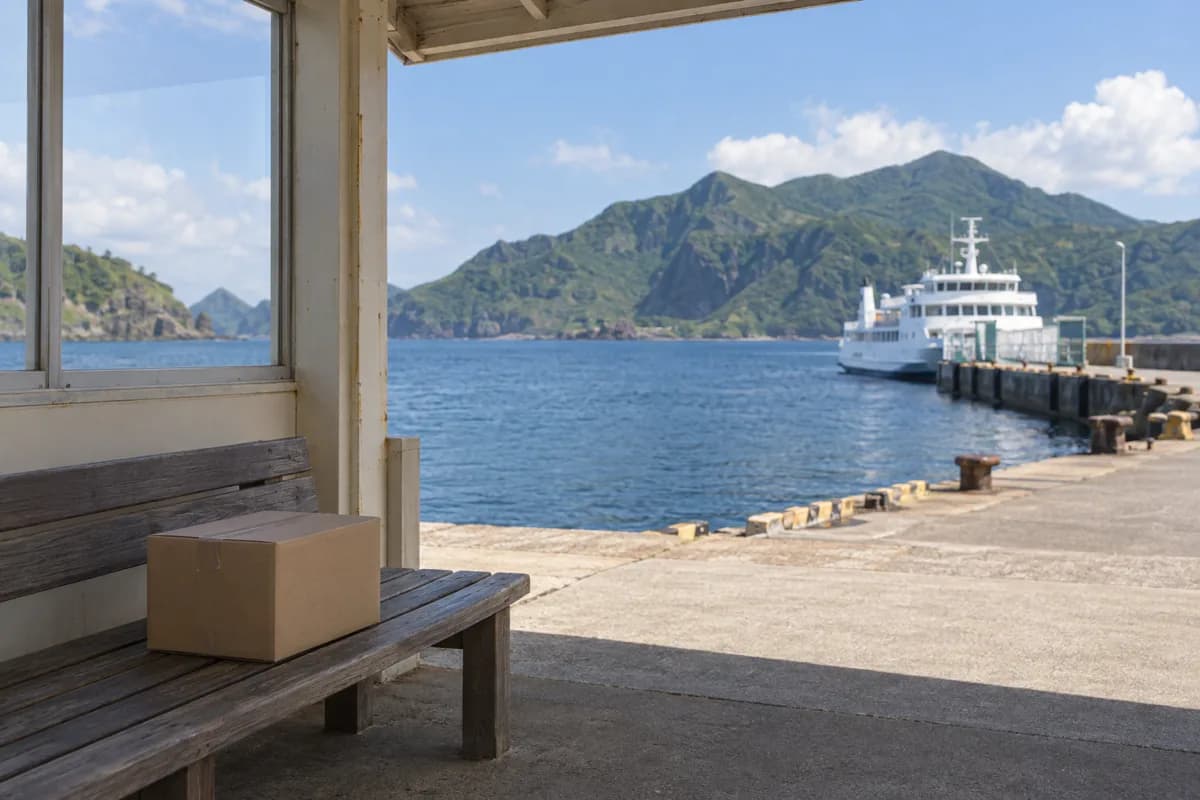島の港の待合所の木のベンチに、無地の段ボール箱がひとつ置かれている。奥には青い海と緑の山、岸壁に停まった白い客船が見える